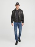 Model draagt een zwarte leren jas, donkergrijze top, blauwe jeans en een pet met 'JA' opdruk.