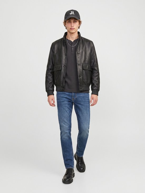 Model draagt een zwarte leren jas, donkergrijze top, blauwe jeans en een pet met 'JA' opdruk.