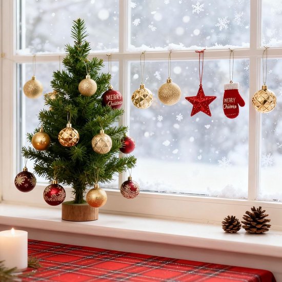 Een kleine kerstboom op een vensterbank met gouden en rode kerstballen, naast een raam met vallende sneeuw.