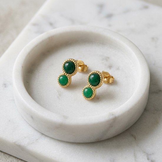 Twee gouden oorknopjes met groene stenen geplaatst in een wit marmeren bakje.