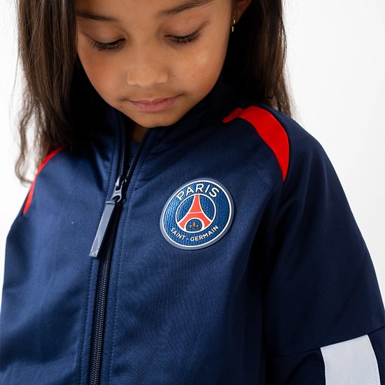Close-up van het marineblauwe PSG trainingsjack met het Paris Saint-Germain logo op de borst. Het jack heeft rode accenten op de schouders.