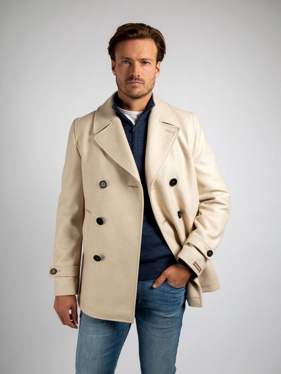 Een beige wollen cabanjas met dubbele knopenrij, gedragen door een man met een blauwe trui en spijkerbroek.