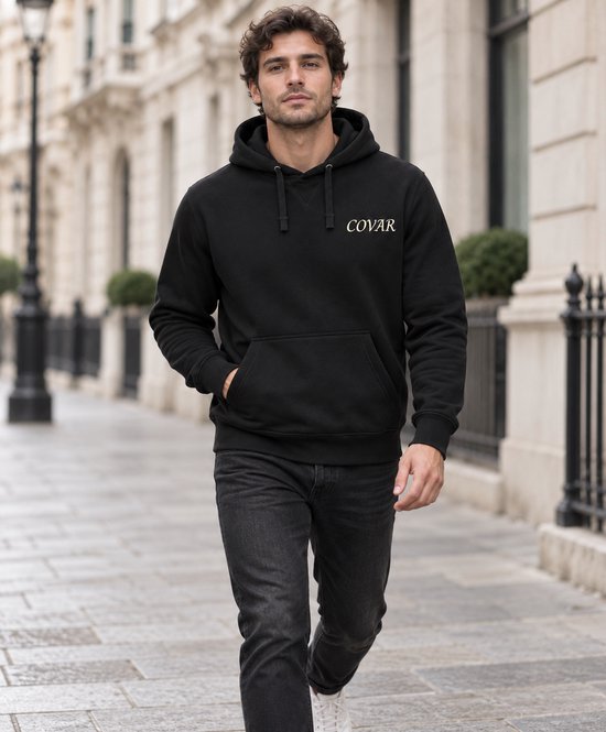 Man met donker haar, gekleed in een zwarte hoodie met COVAR-logo en jeans, wandelt op straat.