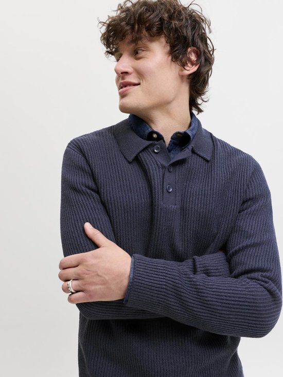 Een man met krullend haar draagt een donkerblauwe gebreide polo met lange mouwen en een denim overhemd eronder.