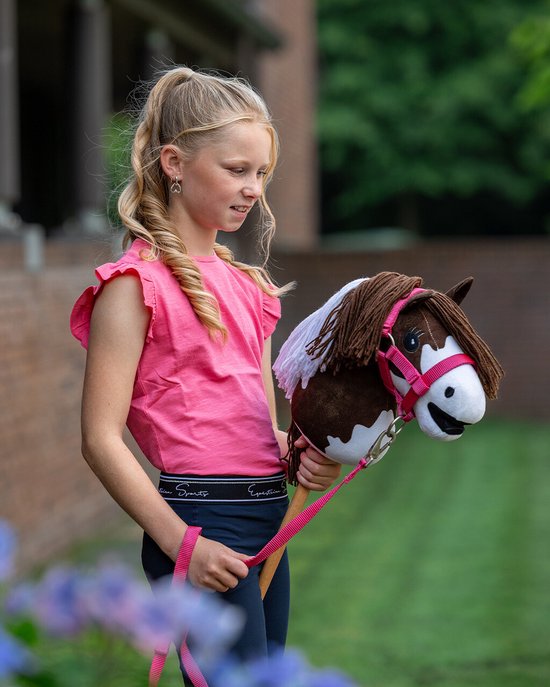 Een meisje met blond haar draagt een roze top en donkerblauwe legging, met een bruin-wit hobby horse met roze hoofdstel.