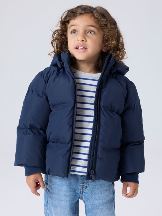 Jongen met krullend haar draagt een donkerblauwe pufferjas met capuchon, gestreept grijs-wit shirt en spijkerbroek.