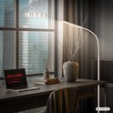 Een beige vloerlamp met gebogen arm, die een bureau met een laptop, boeken en decoratie verlicht bij een raam met uitzicht op de stad.