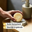 Een hand houdt een oranje shampoobar van Marcel's Green Soap vast boven een zeepbakje.