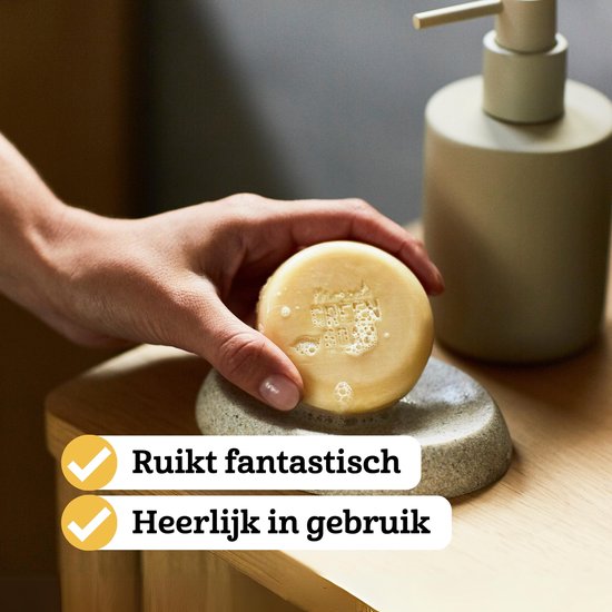 Een hand houdt een oranje shampoobar van Marcel's Green Soap vast boven een zeepbakje.