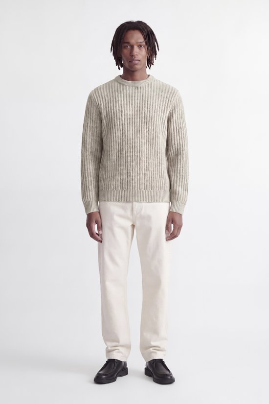 Model in beige grofgebreide trui en witte broek, volledig zicht van voren.
