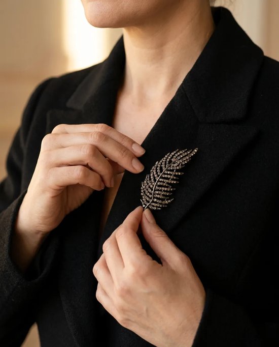 Model draagt een zilverkleurige veer-vormige broche bezet met steentjes op een zwarte blazer.