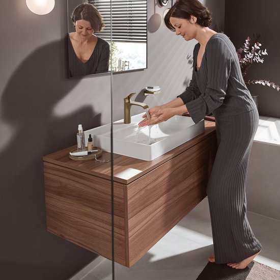 Een vrouw met donker haar wast haar handen bij een witte wastafel met een geborstelde bronzen kraan.