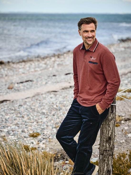 Man op het strand met rood langarm polo shirt met 'Free Spirit' borduring en denim zakje, gedragen met donkere corduroy broek.