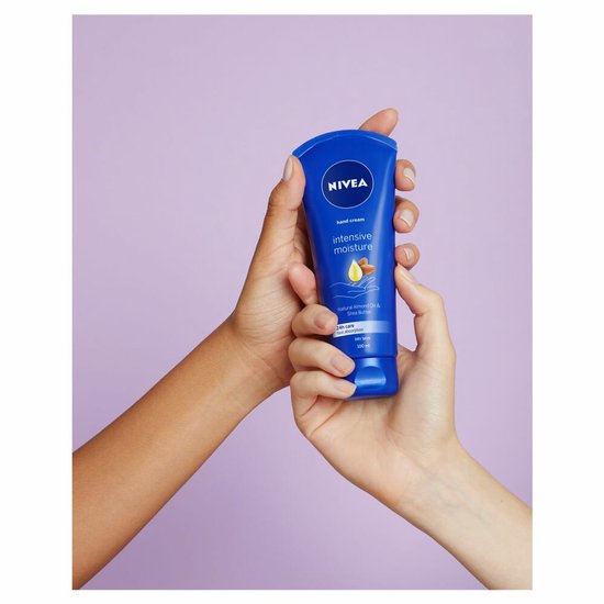 Twee handen houden een blauwe NIVEA handcrème tube met 'Intensive Moisture' erop. De tube toont een druppel en amandelen.