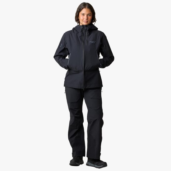 Een vrouw in een zwarte jas en broek, met capuchon op.