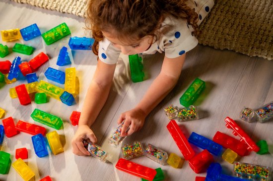 Meisje met krullend haar speelt met rode, blauwe, gele en groene Jelly Blox blokken op de houten vloer.