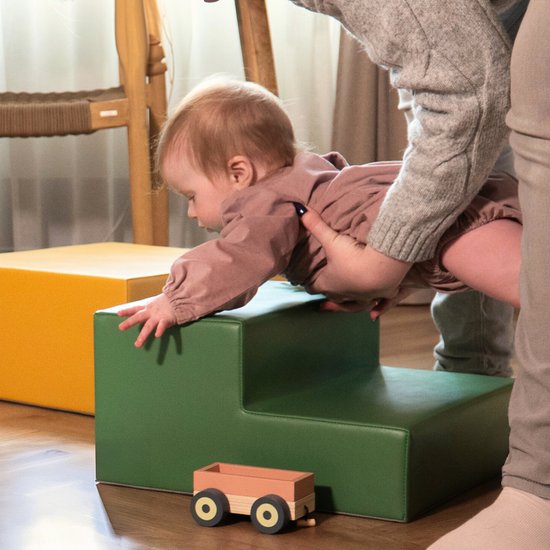 Baby in bruine romper speelt op groene trapeblokken, met gele blokken ernaast.