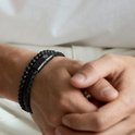 Een zwart gevlochten lederen armband met kralen van zwarte lavasteen en mat zwart, gedragen om een mannenpols.