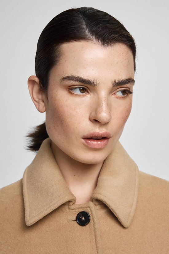 Close-up van een vrouw met sproeten, die een beige jas met een zwarte knoop draagt.