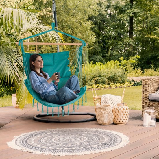 Vrouw met een telefoon in turquoise hangstoel, staand op een metalen standaard op een houten terras.
