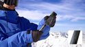 Man in blauwe ski-jas met skibril, houdt een zwarte Insta360 actioncam vast op een verlengstuk. Berglandschap met sneeuw.