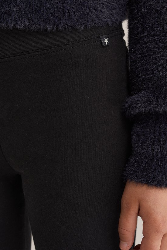 Detail van een zwarte legging met een klein wit sterretje op een zwart label bij de tailleband, naast een donkerblauwe pluizige trui.