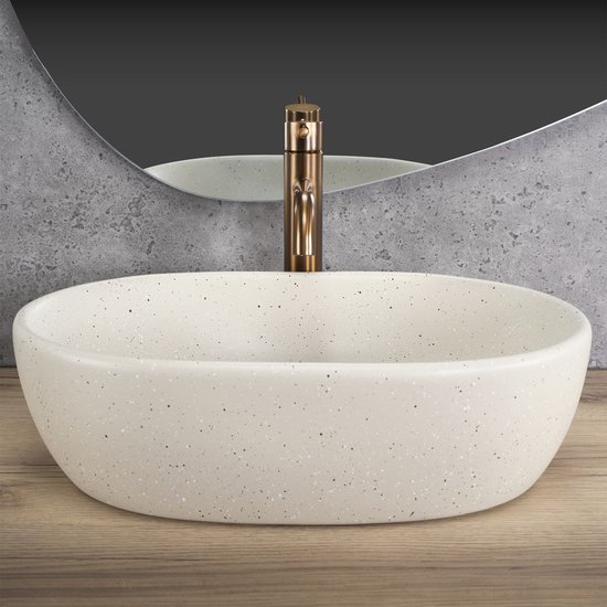 Ovaal beige waskom met spikkels en koperkleurige kraan, met spiegel erboven.