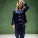 Model draagt een donkerblauwe satijnen pyjama met witte biesjes. De pyjama heeft een knoopsluiting en een borstzakje.