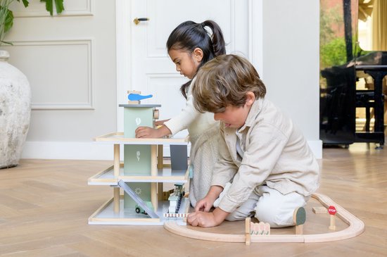 Twee kinderen spelen met een pastelgroene houten speelgoedgarage en een houten treinbaan op een parketvloer.