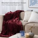 Bordeauxrode woondeken over een beige bank in een woonkamer. Glanzende afwerking.