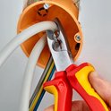 Hand gebruikt rode en gele tang om dikke witte kabel te snijden in een oranje elektriciteitsdoos.