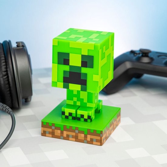 Een felgroene Minecraft Creeper-lamp, staand op een gras- en aarde-voetstuk, met een koptelefoon en controller op de achtergrond.