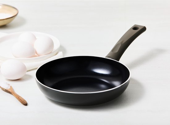 Poêle noire sur une table blanche, avec trois œufs sur une assiette et une cuillère en bois avec du sel.