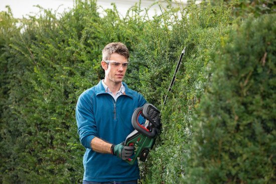 Man met blauwe trui, veiligheidsbril en handschoenen, snoeit een groene heg met heggenschaar.
