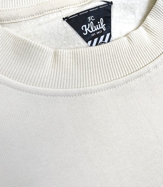Nekdetail van off-white sweater met geribbelde hals en zwart FC Kluif label.