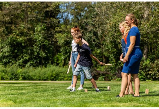 Vier mensen spelen Kubb in een zonnig park; een jongen werpt een stok.
