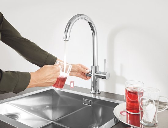 Chroom GROHE kraan die heet water in een glas met theezakje schenkt, met twee mokken ernaast.