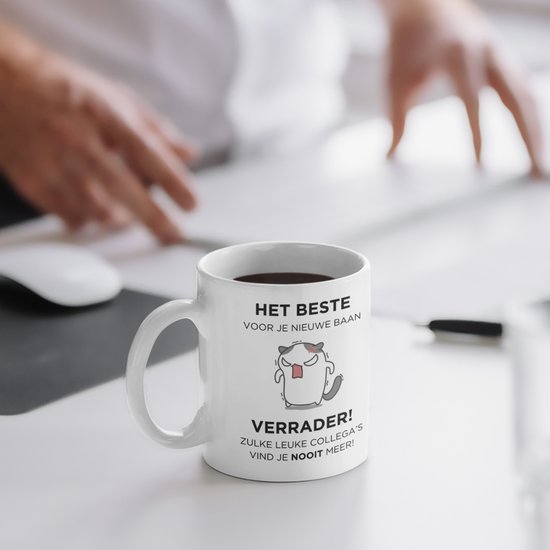 Witte mok met Nederlandse tekst "HET BESTE VOOR JE NIEUWE BAAN VERRADER! ZULKE LEUKE COLLEGA'S VIND JE NOOIT MEER!" en een boos katje, op een bureau met handen op de achtergrond.