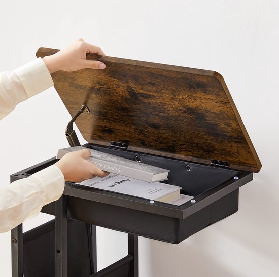 Handen die boeken in het opbergvak van een bijzettafel plaatsen. De tafel heeft een houten blad, een zwarte stoffen mand en een metalen frame.
