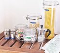 Gecentreerde Dymo Omega labelmaker op een houten tafel met gelabelde voorraadpotten: Pasta, Bonen, Pinda's, Thee, Zout, Peper en Snoep.