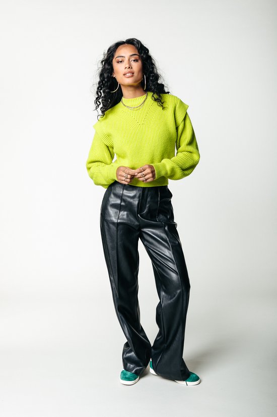 Vrouw in een felgroene trui en zwarte wijde broek, staand.