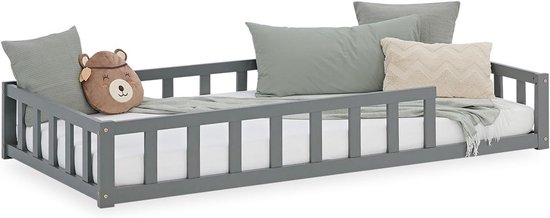 Grijs houten peuterbed met uitvalbeveiliging, opgemaakt met kussens waaronder een berenvormig kussen.