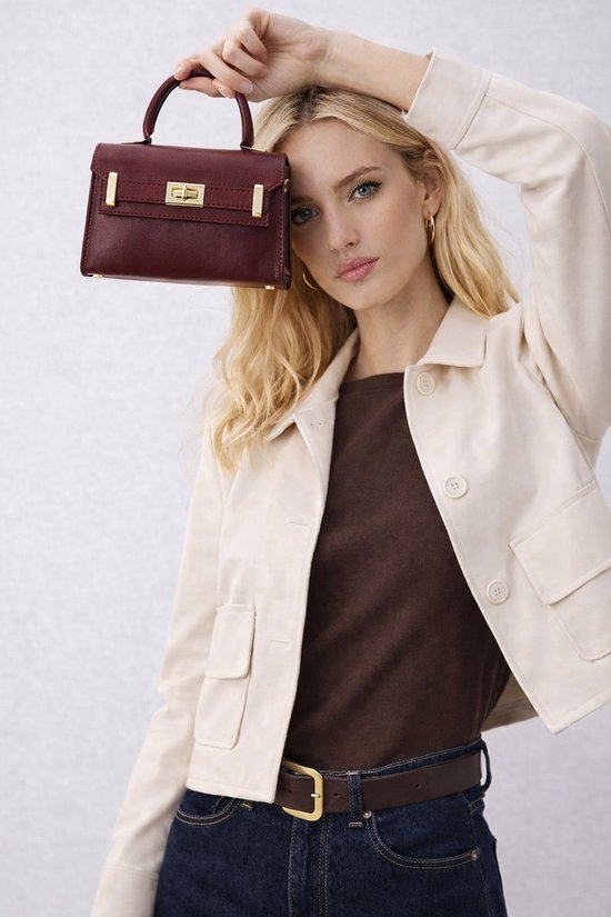 Model houdt bordeauxrode handtas boven haar hoofd, draagt beige blazer, bruine top en jeans.