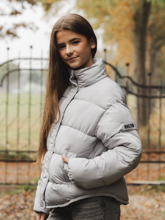 Meisje met lange bruine haren draagt een grijze pufferjas met RVLTN-logo op de mouw.