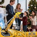 Gele speelgoedkraan wordt als kerstcadeau gegeven aan een jongen door zijn ouders, bij een kerstboom. De kraan heeft een grijze container aan de haak. Een zusje kijkt toe.