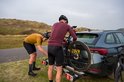 Twee mannen in wielerkleding bevestigen een fiets aan een trekhaakfietsendrager achter een auto, in een duinlandschap.