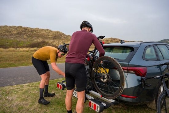Twee mannen in wielerkleding bevestigen een fiets aan een trekhaakfietsendrager achter een auto, in een duinlandschap.