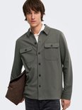 Mannelijk model met olijfgroene, lange mouwen oversized shirt, witte polo eronder en draagt een bruine tas.