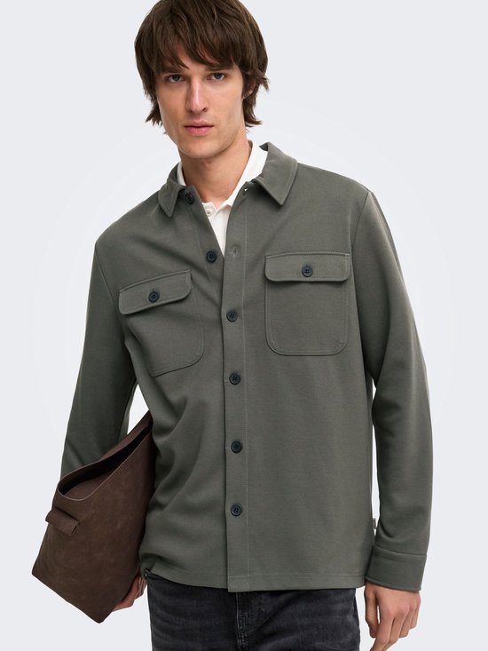 Mannelijk model met olijfgroene, lange mouwen oversized shirt, witte polo eronder en draagt een bruine tas.
