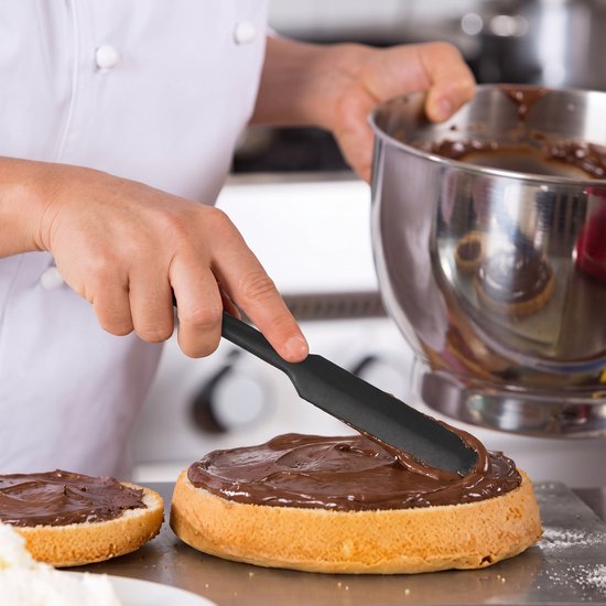 Een hand met een zwarte siliconen spatel smeert chocoladepasta op een cake, naast een metalen mixer.